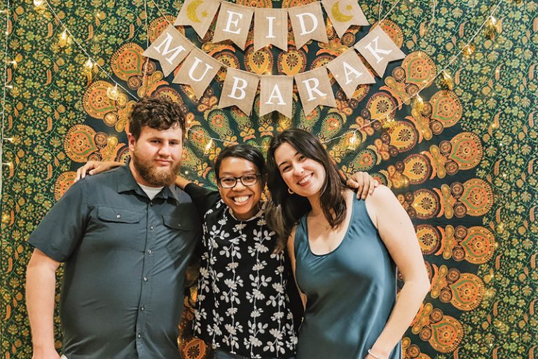 Three students gather in front of an Eid Mubarak sign, which hangs on a tapestry of greens and oranges.