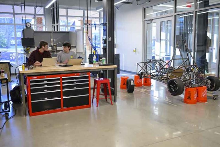 Students discuss the race car they are building in Founders Hall. The shell of the race car is propped up on orange plastic buckets.