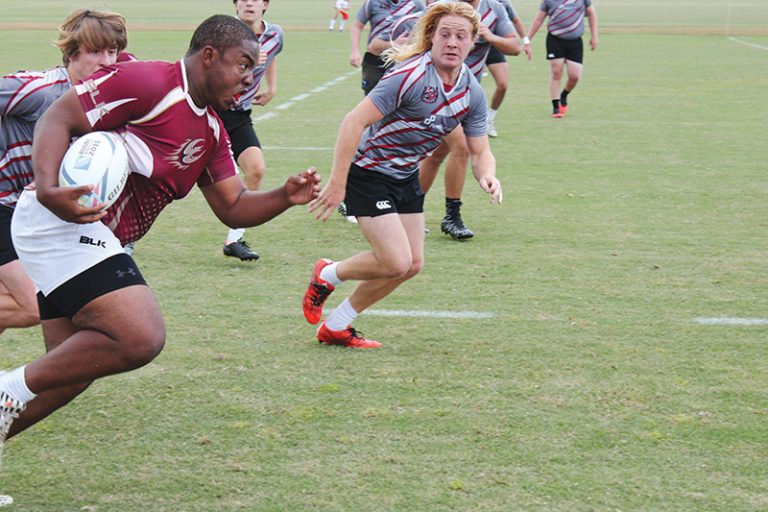 An Elon rugby player dressed in a maroon shirt and white shorts runs with the ball against multiple guys in gray jerseys, black shorts and red cleats.