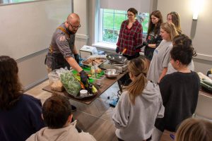 A chef teaches a group of students how to make a meal. He has kale, chicken, spices, vegetables and frying pan on a burner at his table while students gather around.