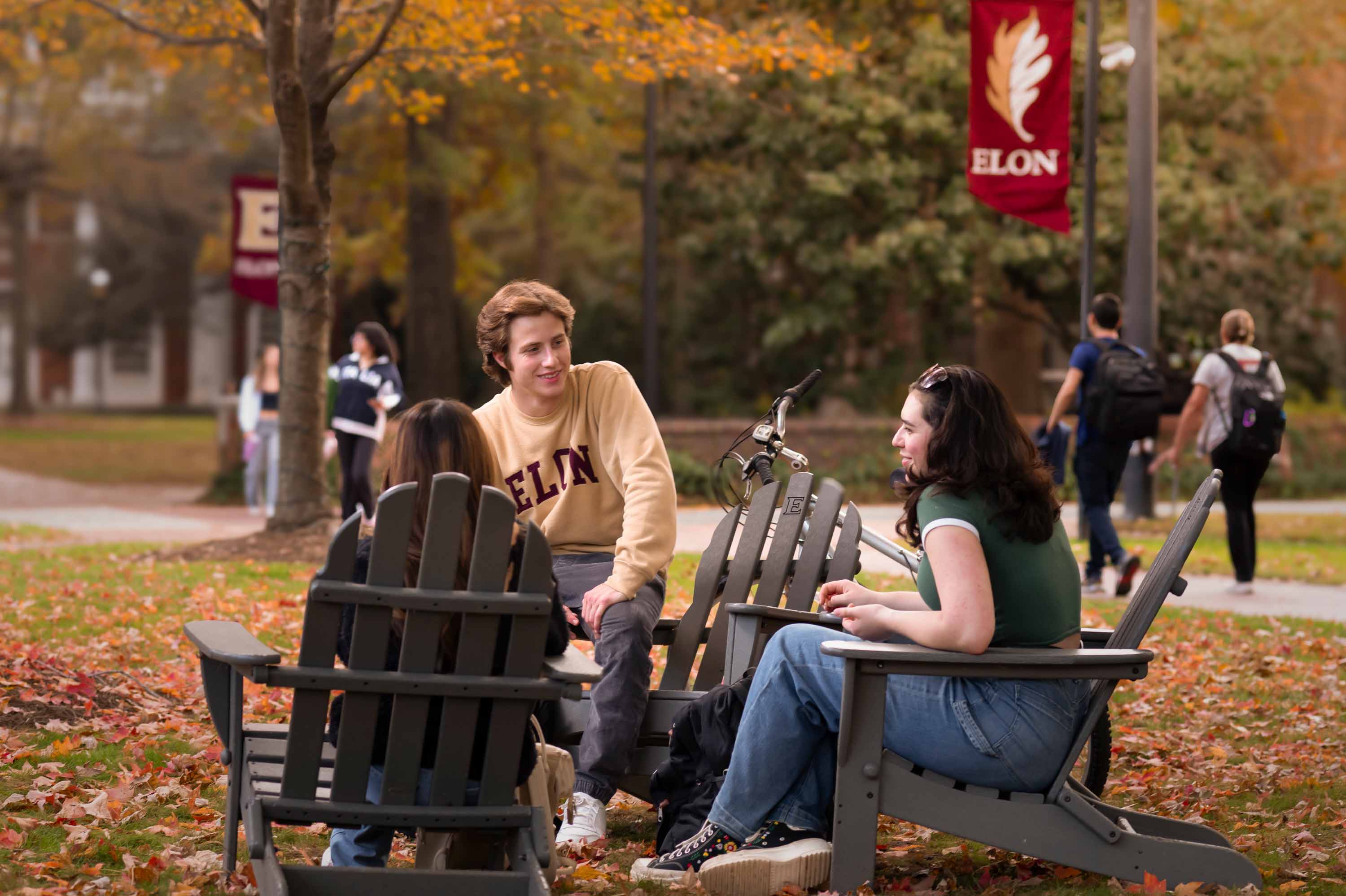 A male and two female students are sitting in Adirondack chairs on campus and talking.