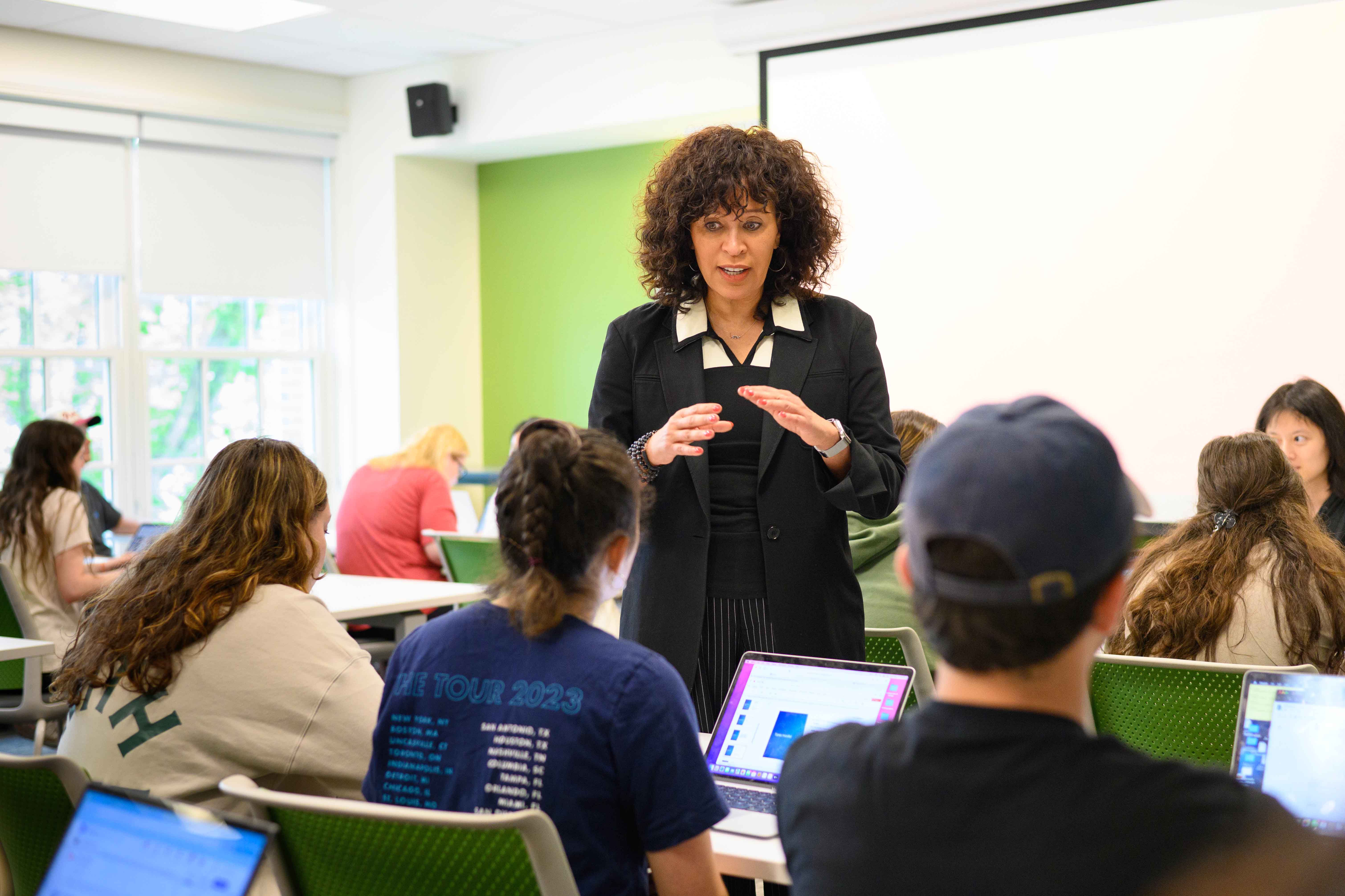 Female professor talks to a group of students around their desks.