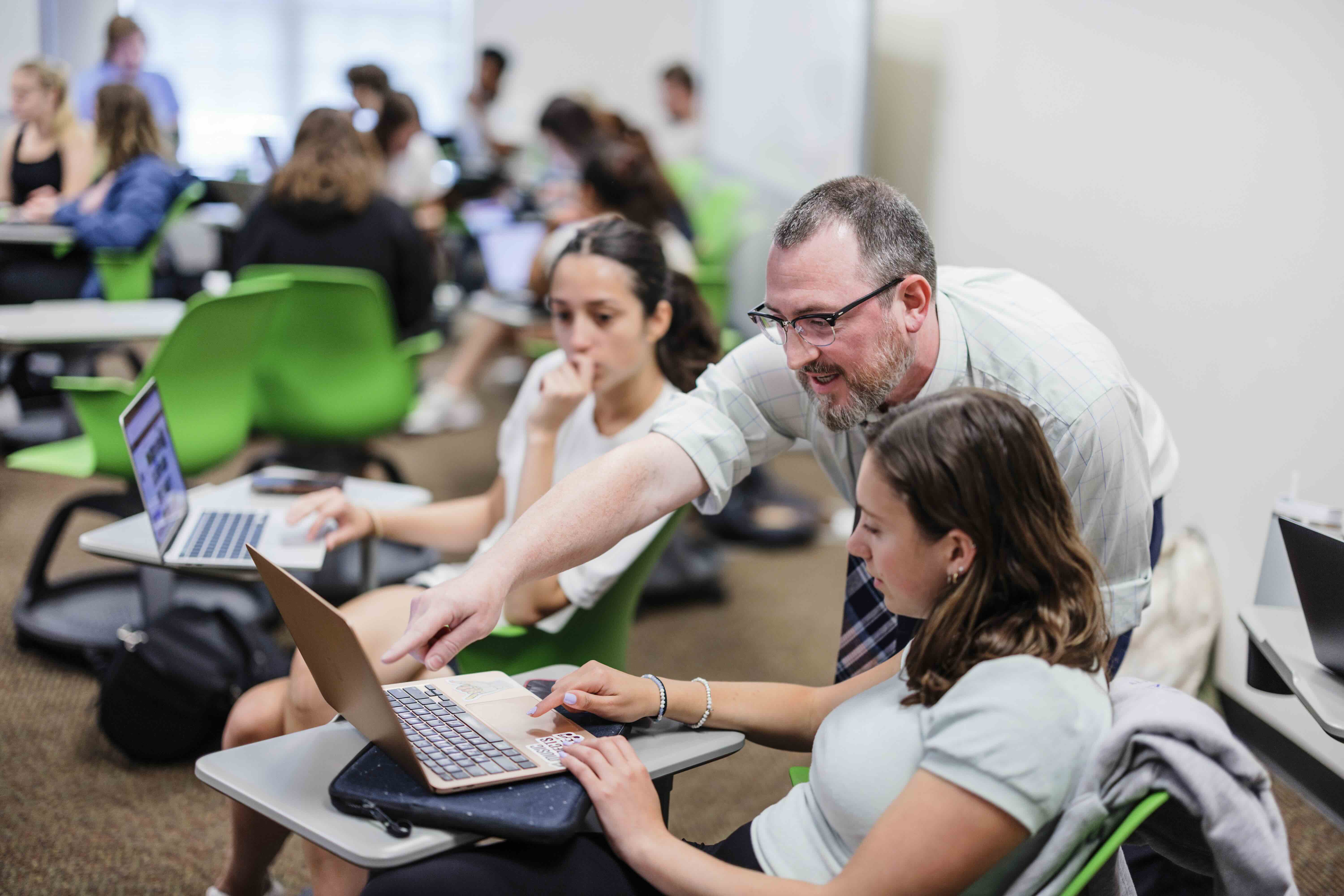 A professor interacts with two students in a classroom.