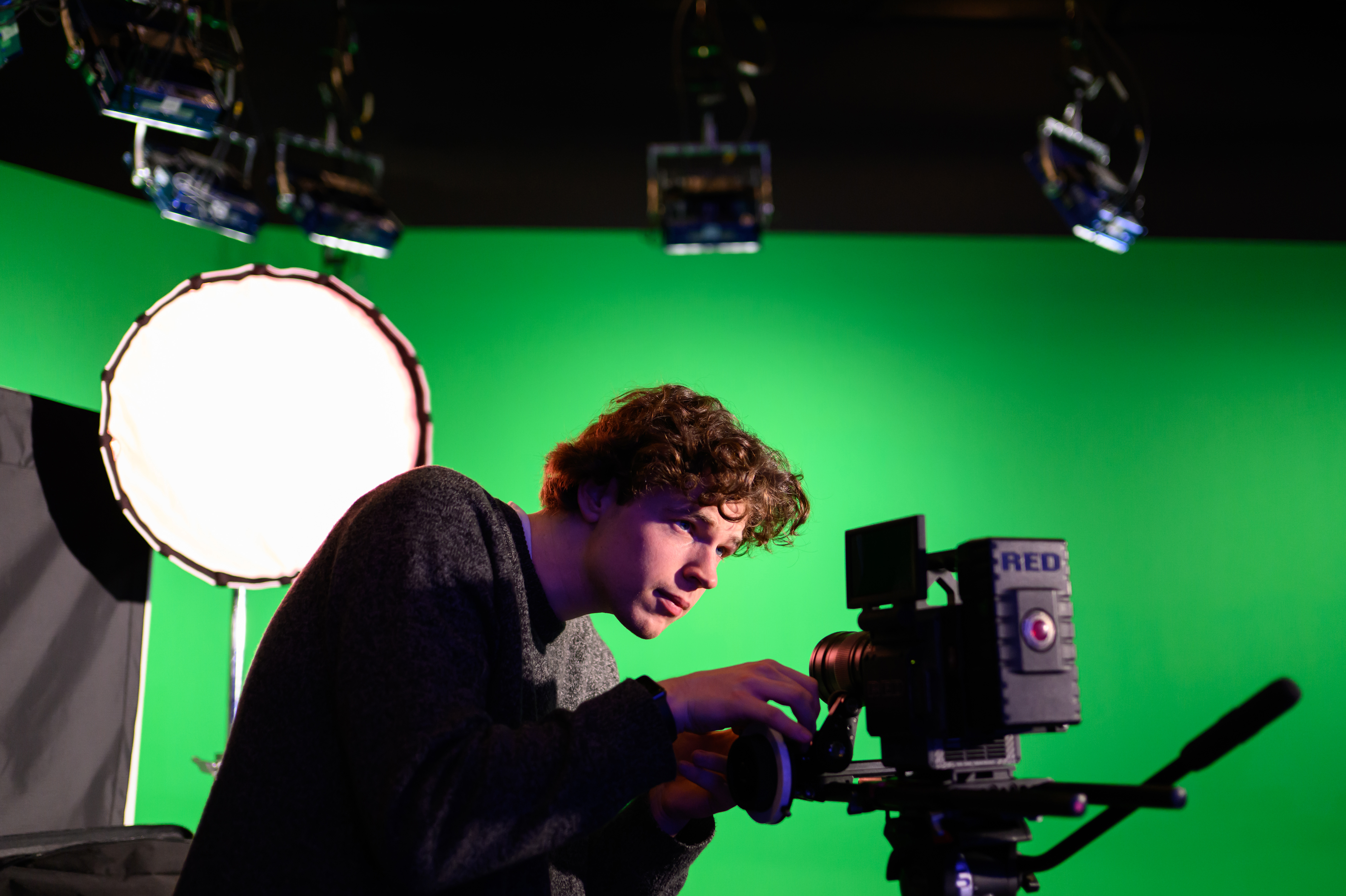 A student with video camera stands in a studio with a green screen.