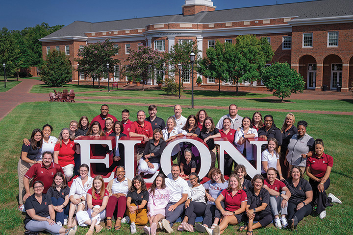 The admissions staff is gathered around the Elon sign on the grass.
