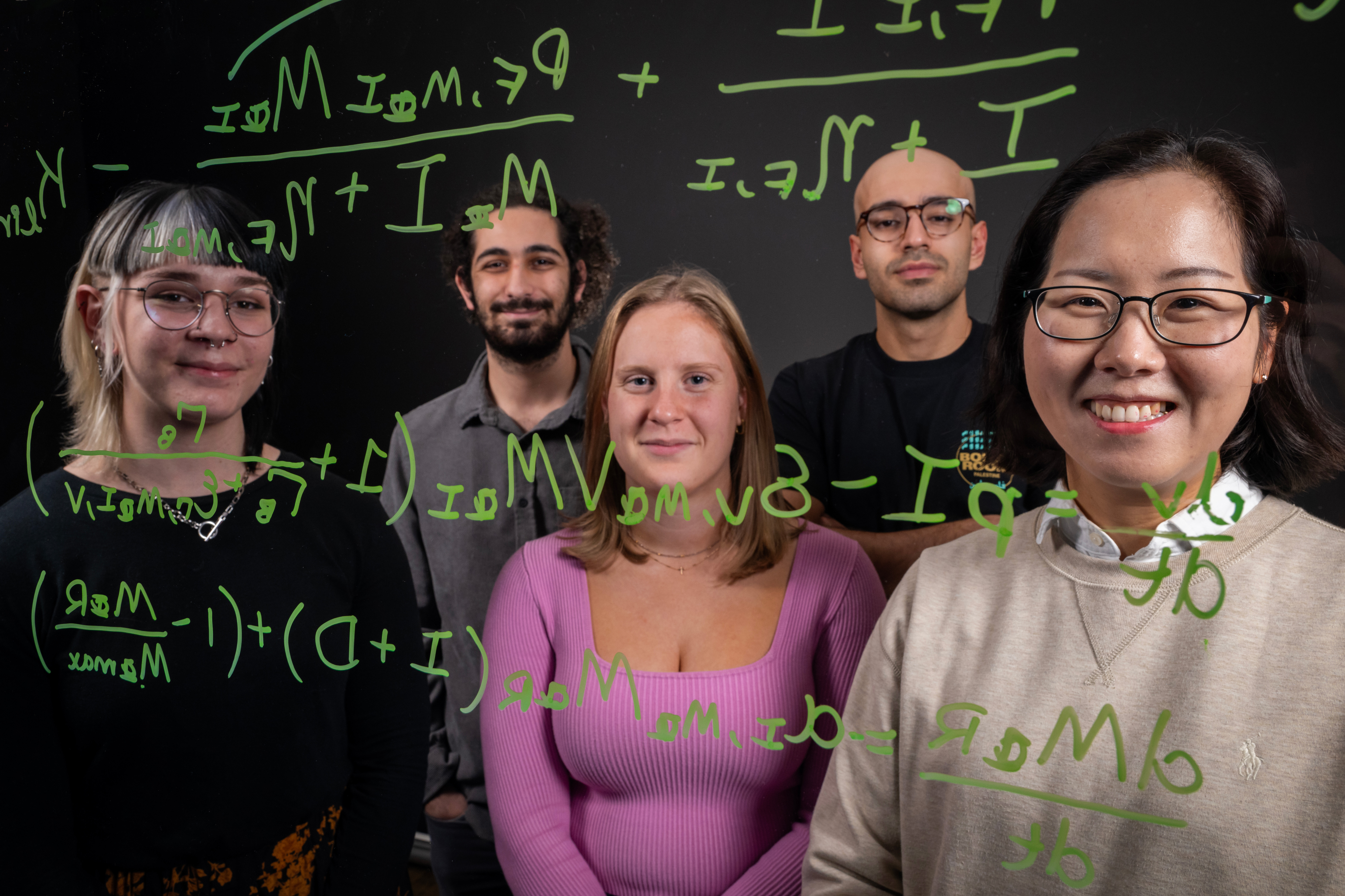Four students stand with a math professor behind a clear white board with math equations on it.