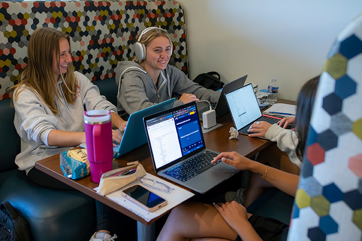 Students are sitting in a booth with their laptops in the Koenigsberger Learning Center. One students is wearing headphones.