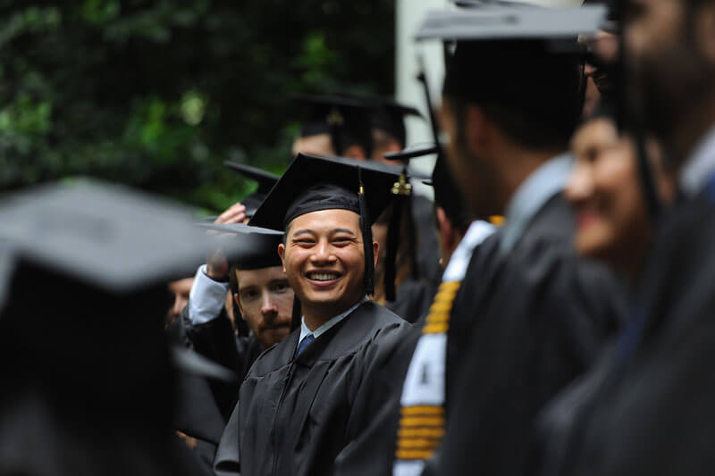 A group of graduates in robes with one single graduate in focus and smiling at the camera.