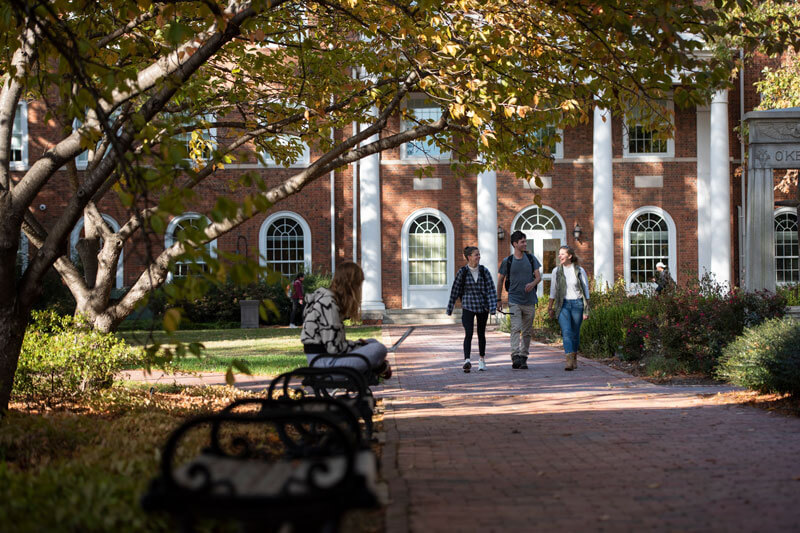 Three students walking along a brick sidewalk in front of a brick building.