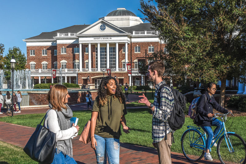 Three students talking in a group in front of the Koury Business Center building.
