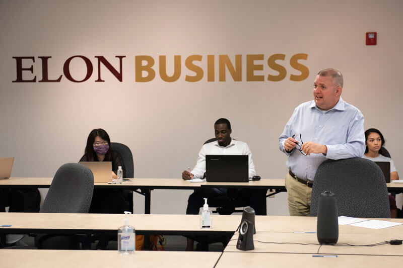 A professor teaching a class in front of a wall with a sign that says Elon Business.