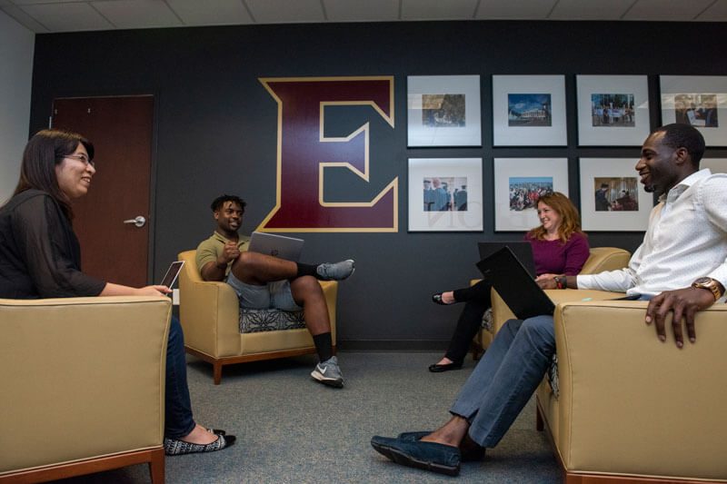 Four students sitting in large, comfy chairs in front of a wall with an Elon E decal and several framed photos.