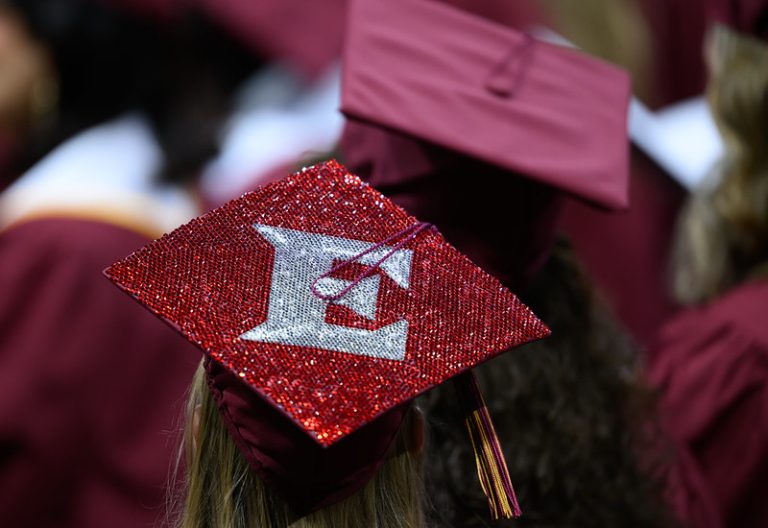 A close-up of a graduation cap decorated with a silver bejeweled letter E in the middle. Red jewels cover the rest of the cap.
