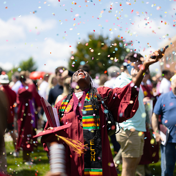 A recent college graduate in a maroon robe smiles as rainbow confetti falls around them