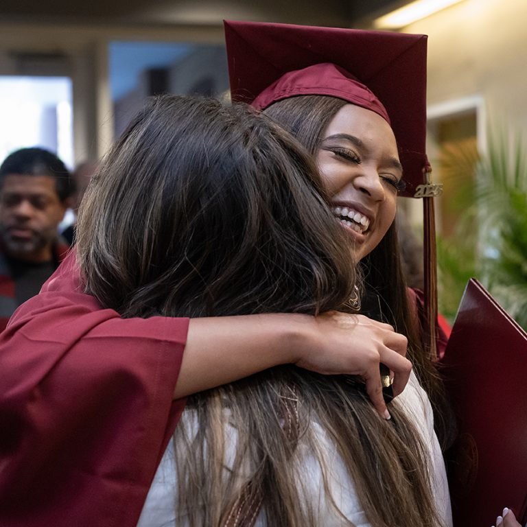 A female graduate wearing a maroon cap and gown holds her diploma and hugs a female loved one.