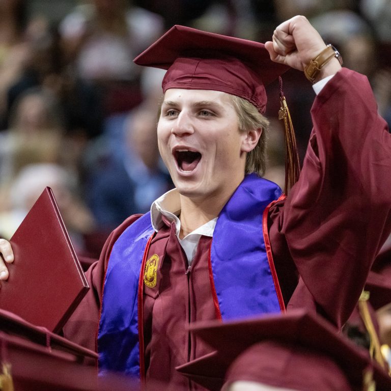 A young man in a graduation cap and gown with his hand up in the air cheering