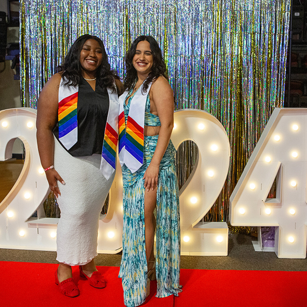 Two students wearing rainbow stoles in front of a lighted 2024 sign