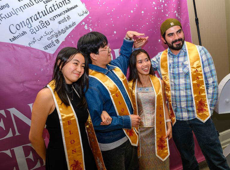 Four students wearing gold Elon stole ceremony stoles pose together and smile in front of a pink ‘Congratulations’ backdrop featuring greetings in multiple languages during the Light the Way API Stole Ceremony.
