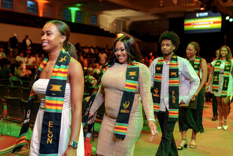Students wearing colorful Kente stoles walk into Elon University’s Donning of the Kente celebration as guests gather in the venue.
