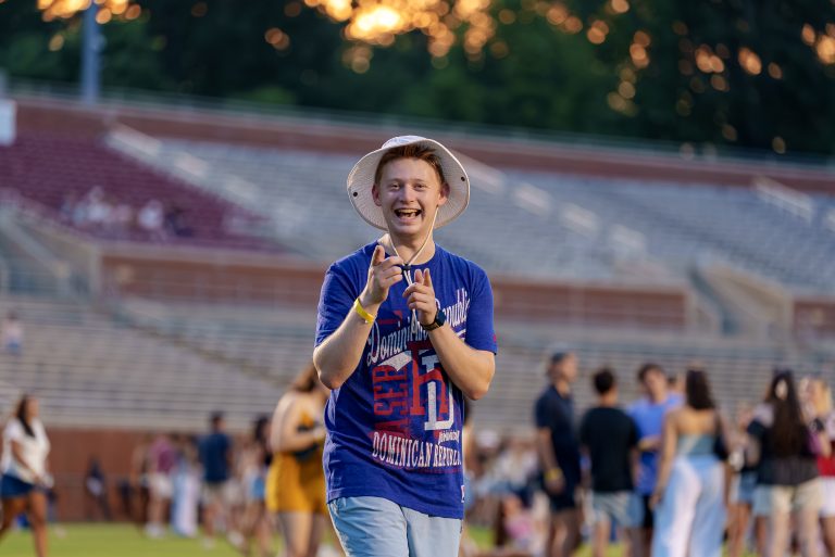 A student wearing a sun hat smiles and points toward the camera during Senior Celebration at Rhodes Stadium.