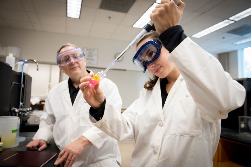 A student wearing goggles and a white lab coat piping liquid into a glass bottle as a professor stands behind them.