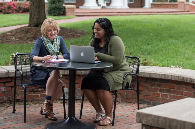 A mentor and their mentee sitting at a round table on a brick patio looking at a laptop and notebook.