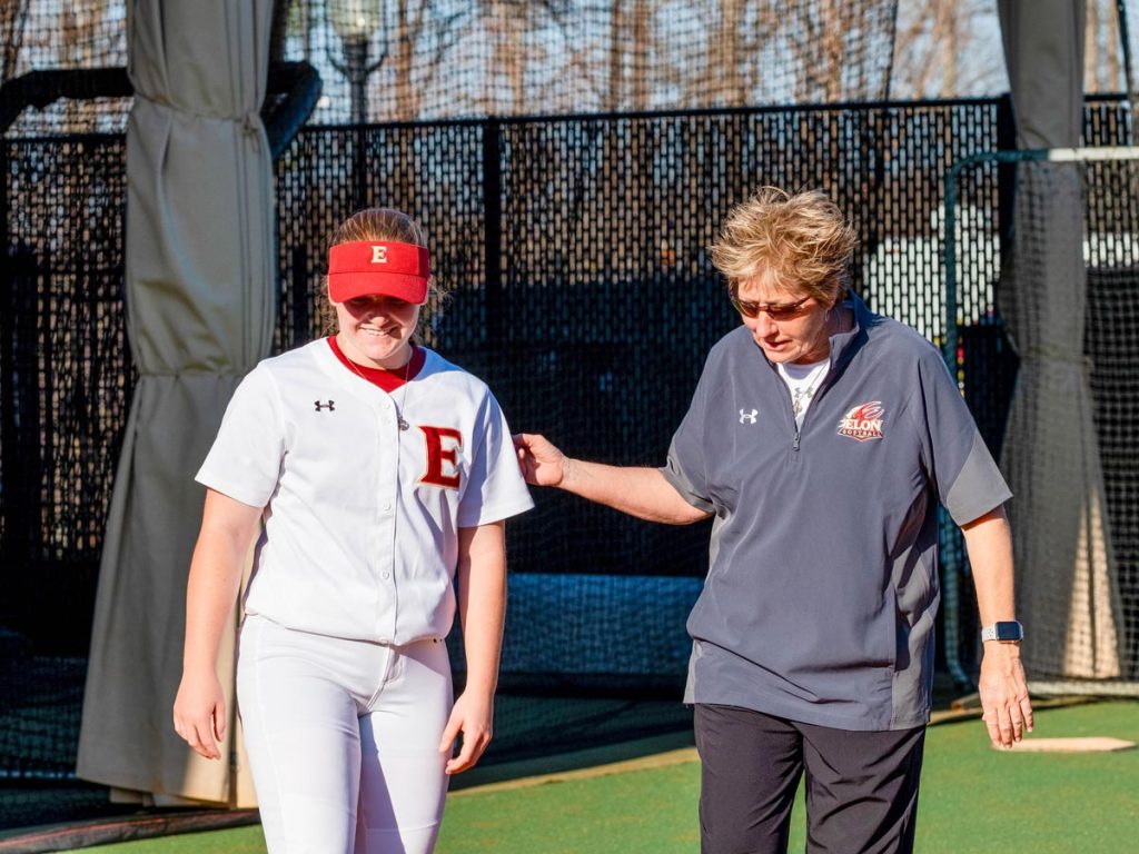 A softball player and softball coach standing in a batting cage area.