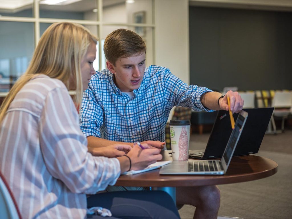 A student tutor and their tutee sitting at a round table looking at a laptop in Elon's walk-in tutoring services center.