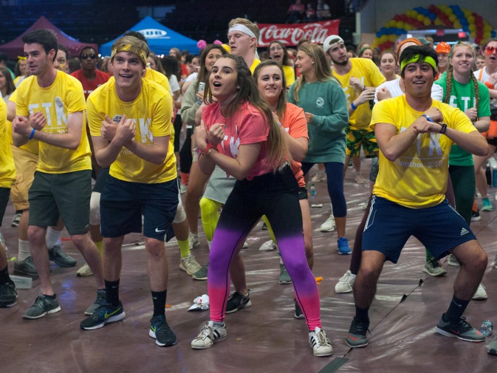 A large group of people wearing brightly colored shirts and outfits dancing in a gym for a charity event.