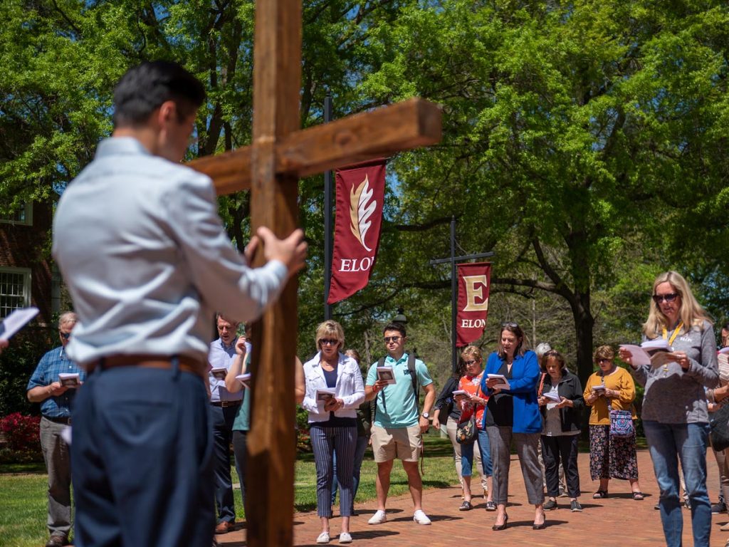 A group of people gathered outside around an individual holding up a large wooden cross for a Stations of the Cross ceremony.