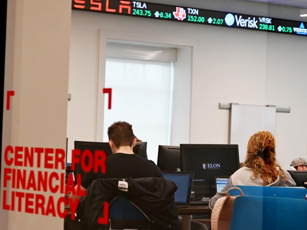 Students working at computers in the Center for Financial Literacy at Elon University, with a digital stock ticker displaying current market prices overhead.