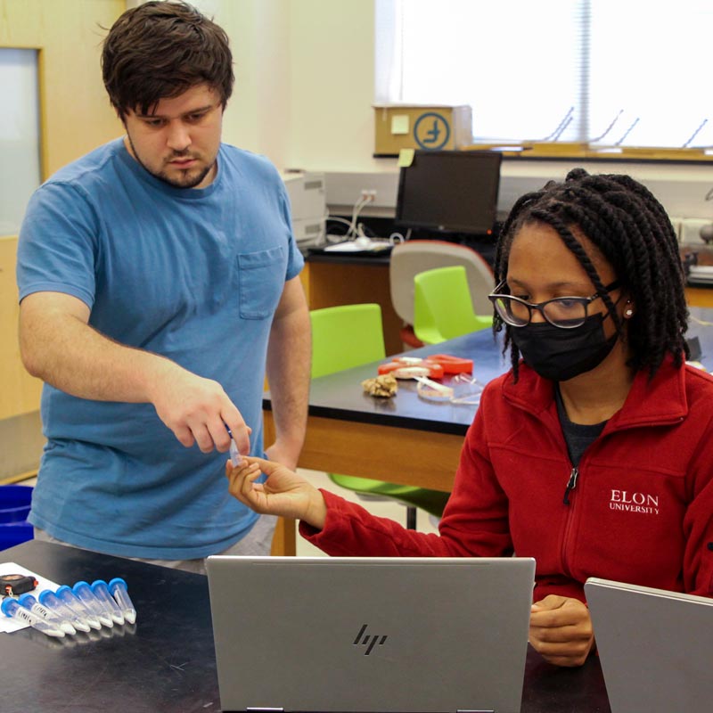 A student standing behind a lab table handing a test tube to another student sitting in front of a laptop.