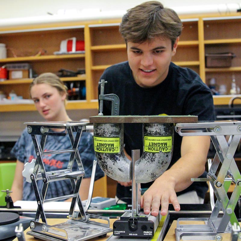 A student working with physics equipment in a lab as another student looks on in the background.