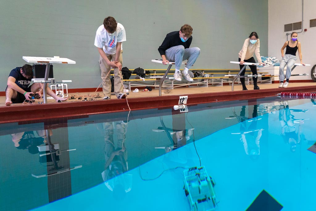 Five students standing by the side of an indoor pool operating underwater vehicles.