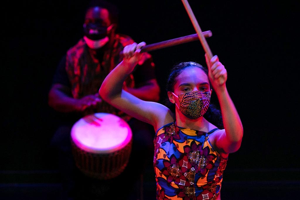 A person holding drumsticks in the air while another person plays a bongo drum in the background.