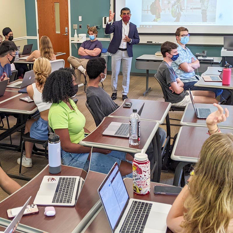 Many students sitting in front of laptops at desks in a classroom while a professor stands in the background.