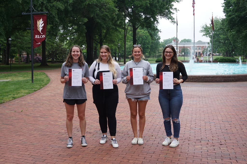 Four Peace Corps students holding up certificates while standing in front of a fountain on Elon's campus.