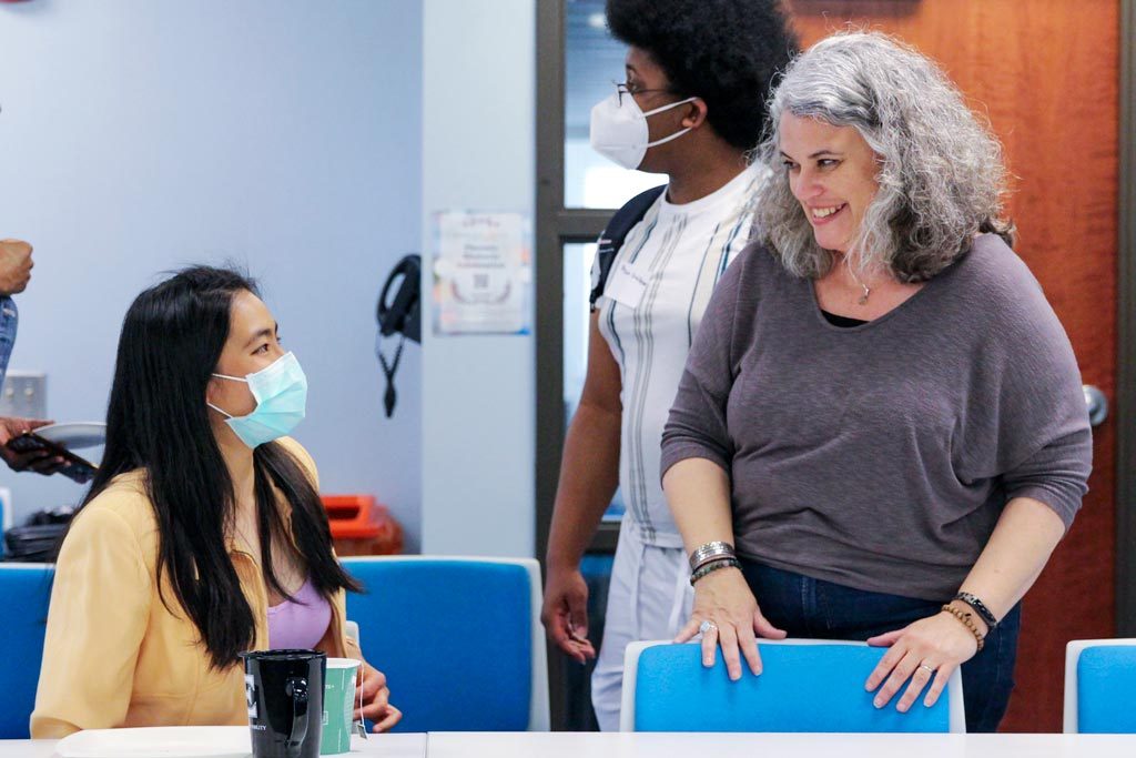 A masked Elon Phoenix Rhetorix student seated at a table talking with a professor standing beside them.