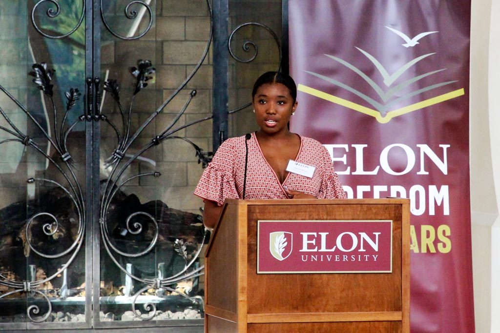 A student giving a speech at a podium with a sign that reads "Elon University" in front of a banner with the Freedom Scholars logo.