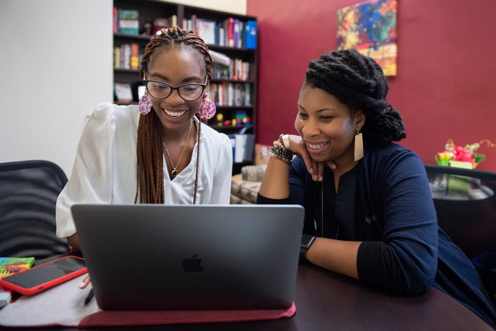 An Elon professor sits at a table in their office looking at a laptop with their student mentee.