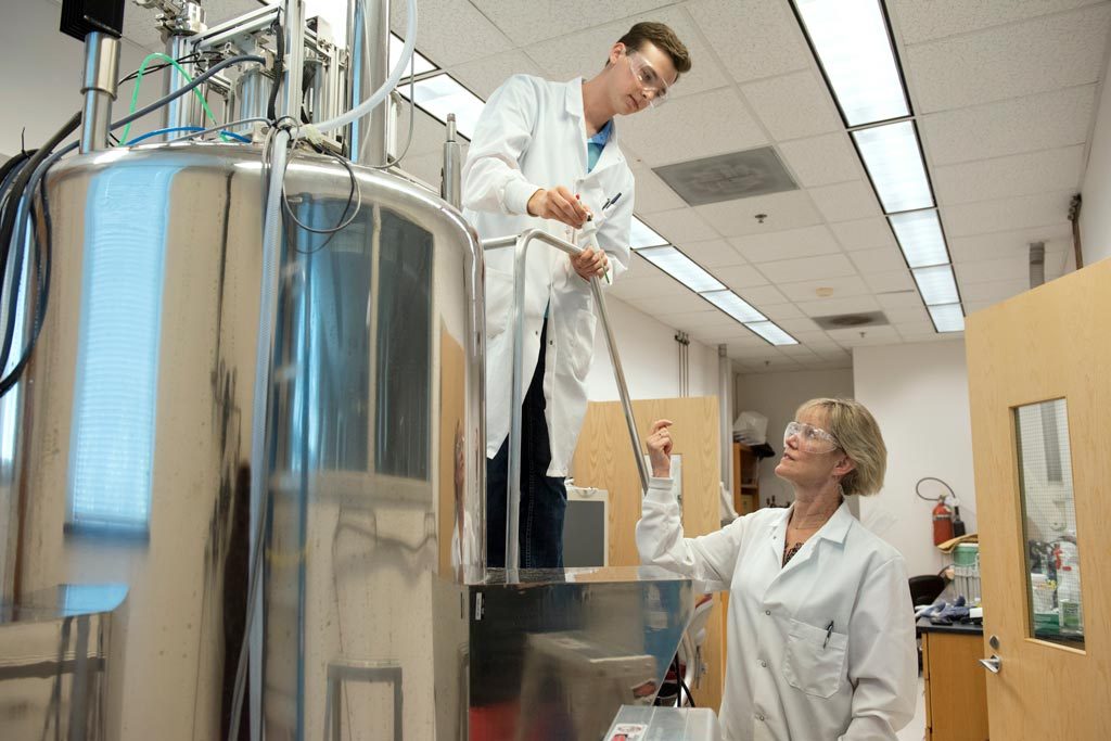 A student wearing goggles and a lab coat standing behind a large silver tank while looking down at their professor.