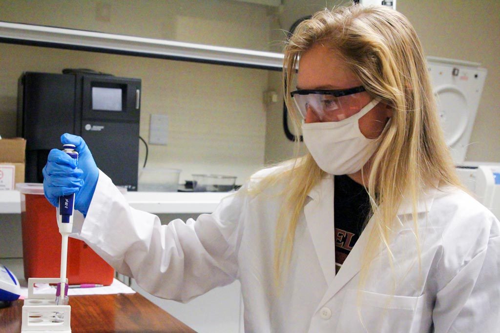 A person wearing a white lab coat with goggles, a mask and blue gloves piping a substance into a test tube.