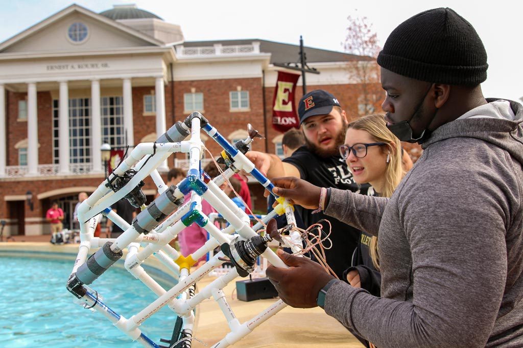 Several students standing around a fountain on Elon's campus while one holds an underwater vehicle made of PVC pipes.