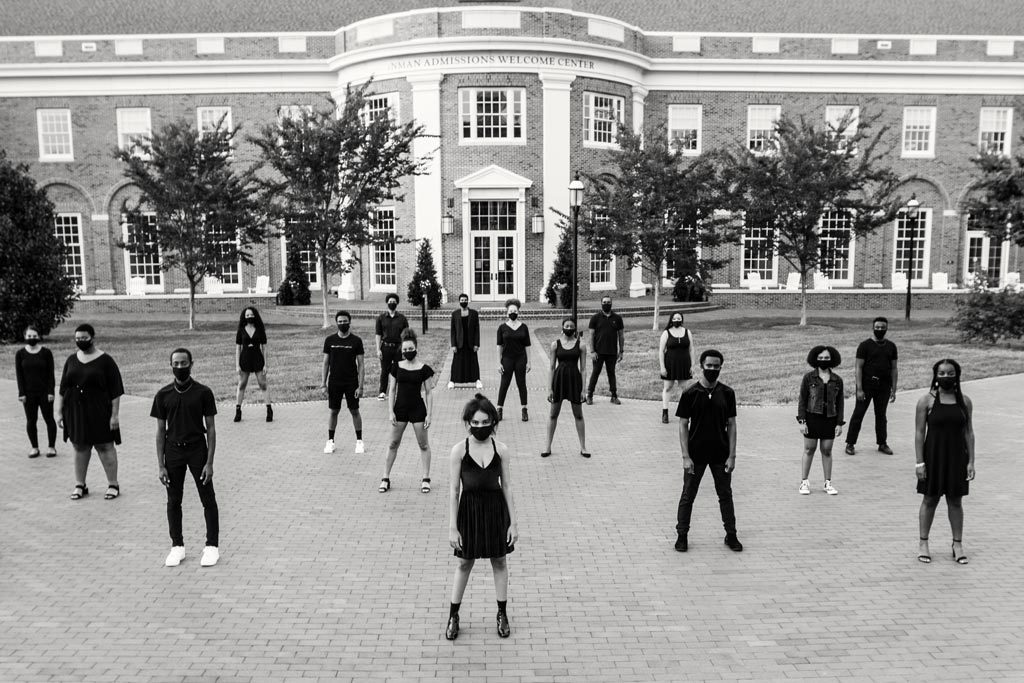 A black and white photograph of the members of all-Black cabaret, “The Moment 2021,” posing in front of Elon's Inman Admissions Welcome Center.