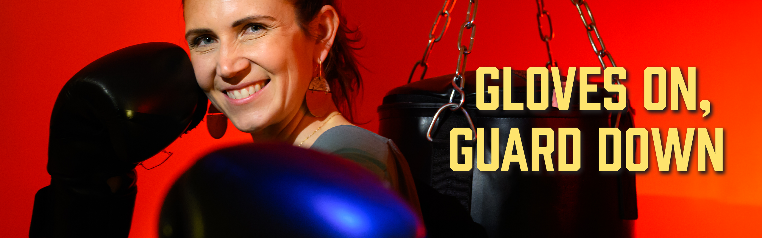A woman wearing boxing gloves smiles beside a hanging punching bag against a bright red background, with text reading “Gloves on, guard down.”