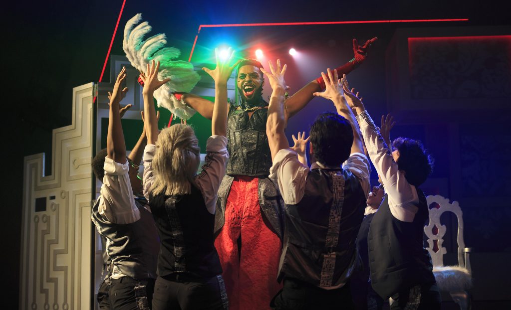 Photo from the Department of Performing Arts production of "We Will Rock You" with actors dancing and singing onstage, circled around an actor holding a fan made of feathers.