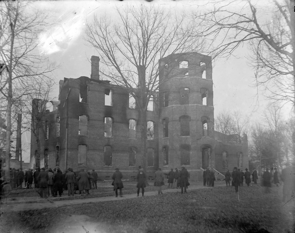 Black and white scan of a historic photograph of campus officials and other spectators view the ruins of the Administration building after the fire.