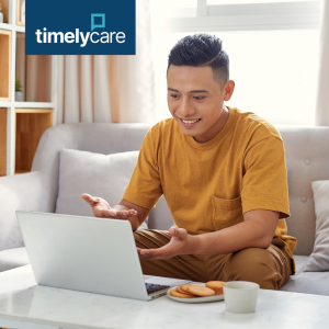 A person sits on a couch, engaged in a video call on a laptop, with the "TimelyCare" logo visible on the screen, in a bright living room setting.
