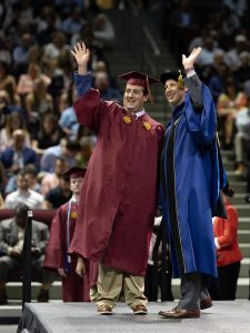 A graduate in maroon regalia and Vice President for Student Life Jon Dooley, in blue academic dress, smile and wave to the crowd during a commencement ceremony on stage.