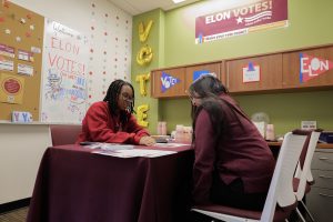 Two students engage in a conversation at a table in the Elon Votes! office, which is decorated with patriotic and voter engagement signage. One student explains materials to the other, promoting civic awareness and participation.